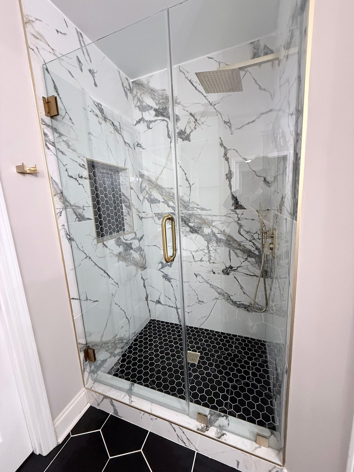 Primary bathroom shower after renovation, floor to ceiling marble panels, glass door, gold fixtures and a black hex tile floor